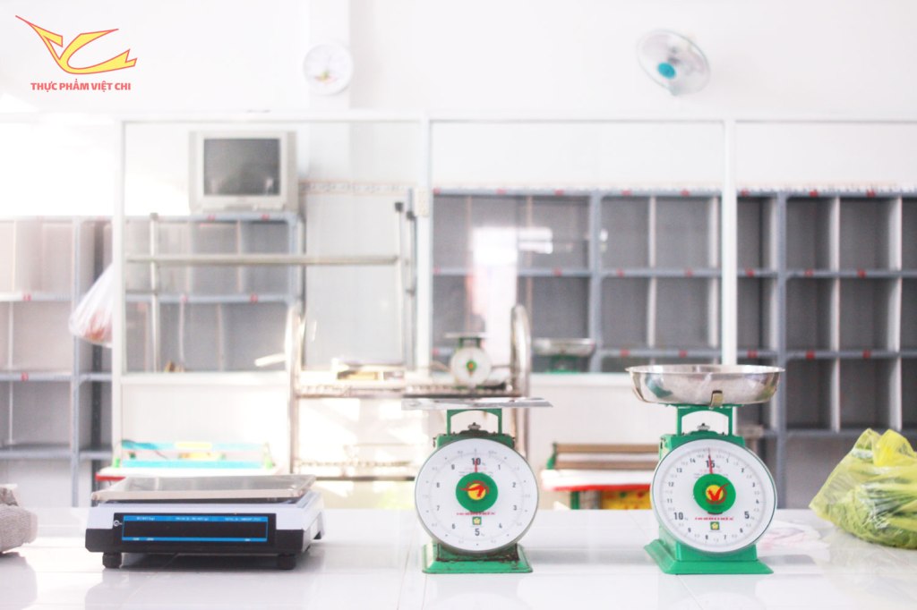 Hai chiếc cân đang đặt trên bàn trong một cửa hàng với kệ trống ở phía sau, có một chiếc tivi và quạt trần.